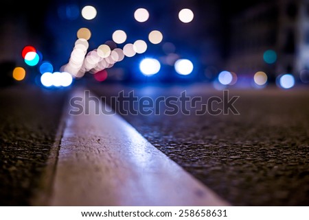 Nights lights of the big city, the night avenue with road markings and headlights of the approaching cars, close up view from asphalt level. In blue tones