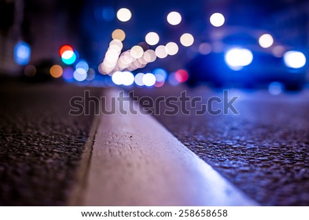 Nights lights of the big city, the night avenue with road markings and headlights of the approaching car, close up view from asphalt level. In blue tones