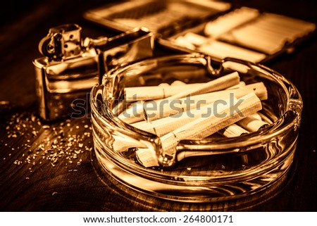 Cigarettes in the ashtray, lighter and cigarette case stands next, on the table broken up tobacco. Focus on the cigarettes, image vignetting and the yellow-orange toning