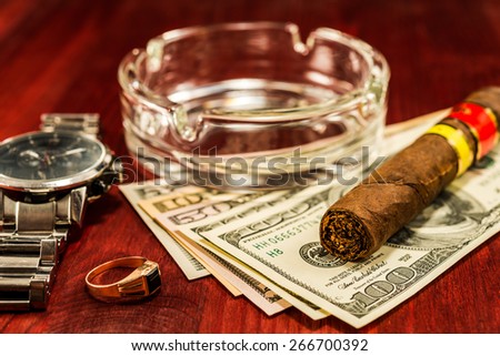 Cuban cigar with glass ashtray on a several dollar bills and gold ring with watches with on the table