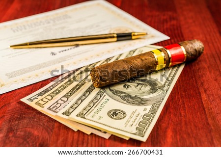 Cuban cigar on a several dollar bills and golden pen with bank check on the table. Focus on the cuban cigar