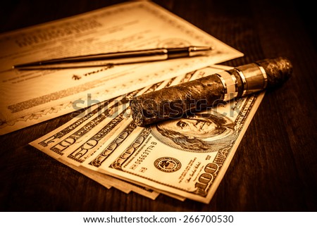 Cuban cigar on a several dollar bills and golden pen with bank check on the table. Focus on the cuban cigar, image vignetting and the yellow-orange toning