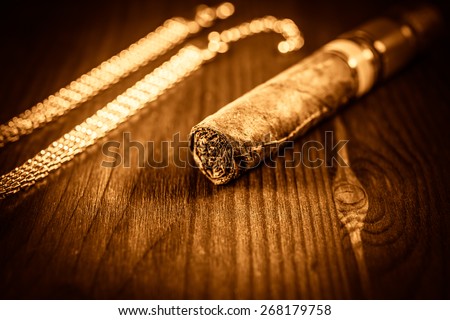 Expensive Cuban cigar and a gold chain on a table in mahogany. De focused image, image vignetting and the yellow-orange toning
