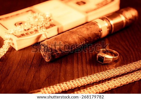 Expensive Cuban cigar and a pack of dollars with gold chain, gold finger ring on a table in mahogany. Image vignetting and the yellow-red toning