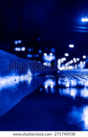 Night city after rain, a reflection of the city at night in the water. View of the stream of cars from the roadside at the asphalt level, image in the blue toning