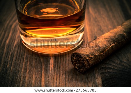 Glass of whiskey and cuban cigar on a wooden table. Focus on the cuban cigar, image vignetting and the orange-blue toning