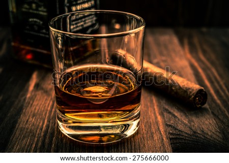 Bottle of whiskey and a glass with cuban cigar on a wooden table. Angle view, image vignetting and the orange-blue toning