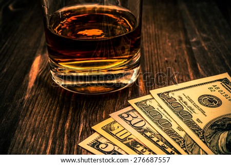 Glass of whiskey with money on a wooden table. Close up view, focus on the money, image vignetting and the orange-blue toning