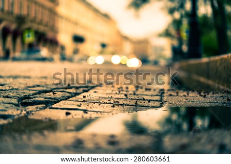 City central square paved with stone after a rain, headlights from cars in the distance. View from the pavement level next to the roadside puddle, image in the orange-blue toning