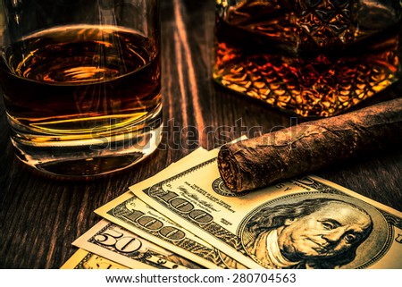 Decanter of whiskey and a glass with cuban cigar and money on a wooden table. Angle view, focus on the cuban cigar, image vignetting and the orange-blue toning