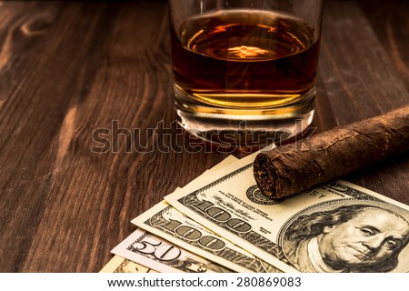 Glass of whiskey and a money with cuban cigar on a wooden table. Close up view, focus on the cuban cigar
