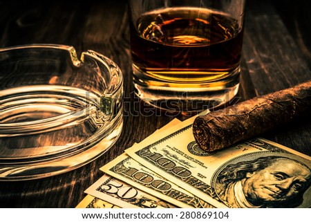 Glass of whiskey and a money with cuban cigar on a wooden table. Close up view, focus on the cuban cigar, image vignetting and the orange-blue toning