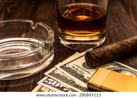 Glass of whiskey and a money with cuban cigar and golden lighter on a wooden table. Close up view, focus on the cuban cigar
