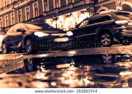 The bright lights of the evening city, driving cars. View from the pavement level next to the roadside puddle, image in the orange-blue toning
