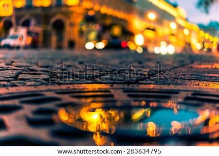 The bright lights of the evening city after rain, headlights from cars in the distance. View from the hatch on the pavement level, image in the orange-blue toning