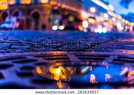 The bright lights of the evening city after rain, headlights from cars in the distance. View from the hatch on the pavement level, image in blue tones