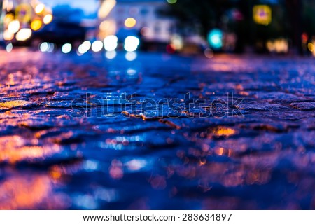 The bright lights of the evening city after rain, headlights from cars in the distance. View from the pavement level, image in blue tones