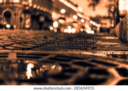 The bright lights of the evening city after rain, headlights from cars in the distance. View from the hatch on the pavement level, image in the orange-purple toning