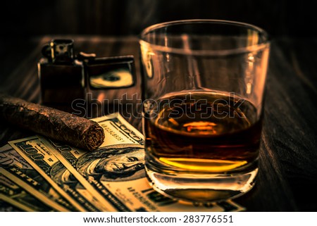 Glass of whiskey and a money with cuban cigar and lighter on a wooden table. Angle view, focus on the cuban cigar, image vignetting and the orange-blue toning