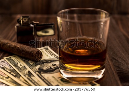 Glass of whiskey and a money with cuban cigar and lighter on a wooden table. Angle view, focus on the cuban cigar
