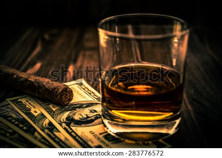 Glass of whiskey and a money with cuban cigar on a wooden table. Angle view, focus on the cuban cigar, image vignetting and the orange-blue toning