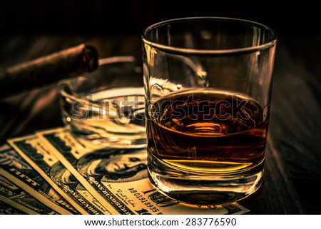 Glass of whiskey and a money with cuban cigar on a wooden table. Angle view, shallow depth of field, focus on the glass of whiskey, image vignetting and the orange-blue toning
