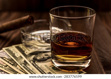 Glass of whiskey and a money with cuban cigar on a wooden table. Angle view, shallow depth of field, focus on the glass of whiskey