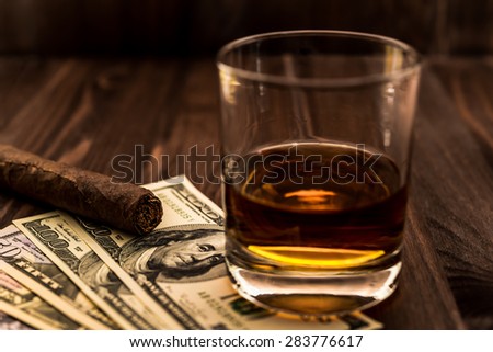 Glass of whiskey and a money with cuban cigar on a wooden table. Angle view, focus on the cuban cigar