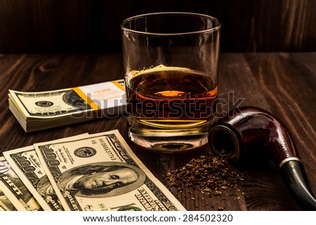 Glass of whiskey with a money and tobacco pipe with tobacco leaves are scattered on the wooden table. Angle view, focus on the tobacco pipe