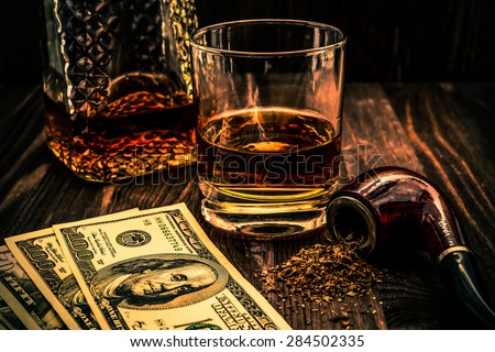 Glass of whiskey with a money and tobacco pipe with tobacco leaves are scattered on the wooden table. Angle view, focus on the tobacco pipe, image vignetting and the orange-blue toning