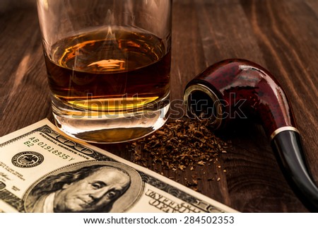 Glass of whiskey with a money and tobacco pipe with tobacco leaves are scattered on the wooden table