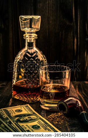 Glass of whiskey with a money and tobacco pipe with tobacco leaves are scattered on the wooden table. Angle view, focus on the tobacco pipe, image vignetting and the orange-blue toning