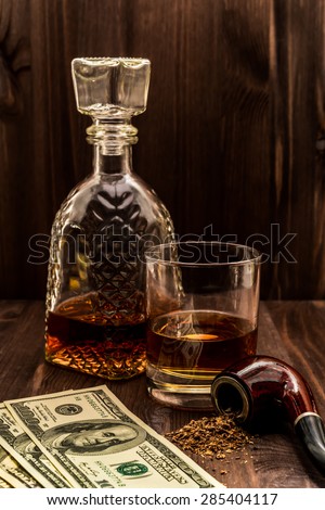 Glass of whiskey with a money and tobacco pipe with tobacco leaves are scattered on the wooden table. Angle view, focus on the tobacco pipe