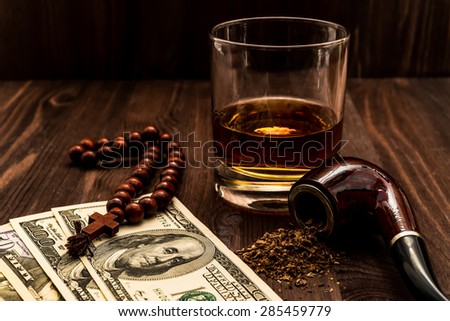 Glass of whiskey and rosary with cross and a money with tobacco pipe on the wooden table. Angle view, focus on the tobacco pipe