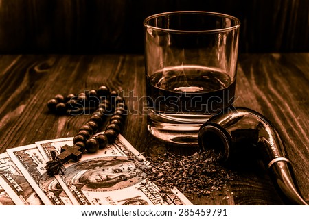 Glass of whiskey and rosary with cross and a money with tobacco pipe on the wooden table. Angle view, focus on the tobacco pipe, image in the orange toning