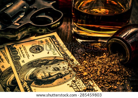 Glass of whiskey and revolver with a money and tobacco pipe on the wooden table. Image vignetting and the orange-blue toning
