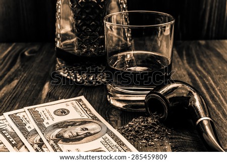 Glass of whiskey with a money and tobacco pipe with tobacco leaves are scattered on the wooden table. Angle view, focus on the tobacco pipe, image in the yellow toning