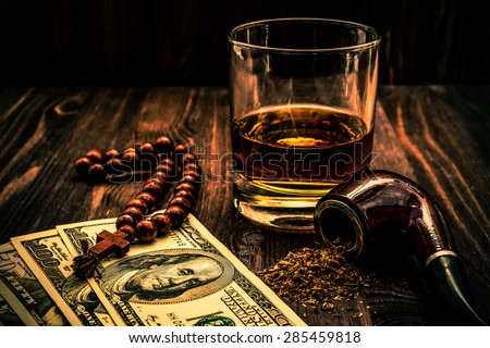 Glass of whiskey and rosary with cross and a money with tobacco pipe on the wooden table. Angle view, focus on the tobacco pipe, image vignetting and the orange-blue toning