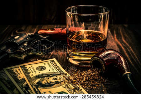 Glass of whiskey and revolver with a money and tobacco pipe on the wooden table. Angle view, focus on the tobacco pipe, image vignetting and the orange-blue toning
