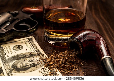 Glass of whiskey and revolver with a money and tobacco pipe on the wooden table