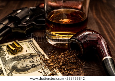 Glass of whiskey and revolver with a money and tobacco pipe on the wooden table