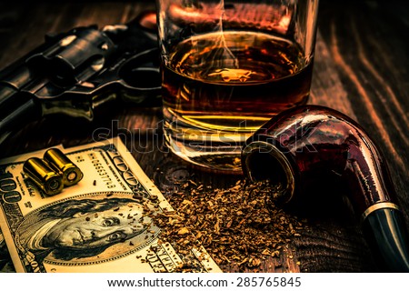 Glass of whiskey and revolver with a money and tobacco pipe on the wooden table. Image vignetting and the orange-blue toning