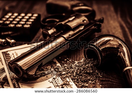 Money and a gun with a box of ammunition with a pipe on a wooden table. Image vignetting and the yellow-orange toning