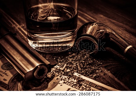 Glass of whiskey and revolver with a money and tobacco pipe on the wooden table. Focus on the tobacco pipe. Image vignetting and the yellow-orange toning