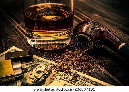 Glass of whiskey with a money and tobacco pipe with tobacco leaves are scattered on the wooden table. Focus on the tobacco pipe, image vignetting and the orange-blue toning