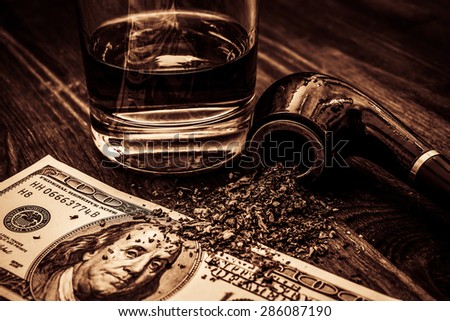 Glass of whiskey with a money and tobacco pipe with tobacco leaves are scattered on the wooden table. Focus on the tobacco pipe, image vignetting and the yellow-orange toning