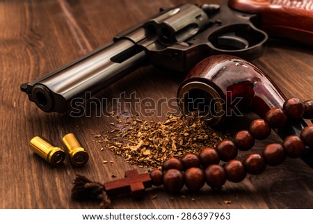 Revolver and a rosary with cross and tobacco pipe on the wooden table. Focus on the tobacco pipe