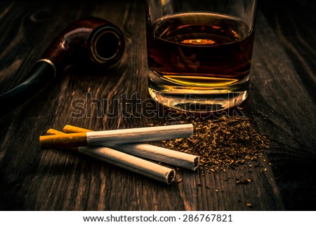 Glass of whiskey and three cigarettes with tobacco leaves and pipe on a wooden table. Focus on the tobacco, image vignetting and the orange-blue toning