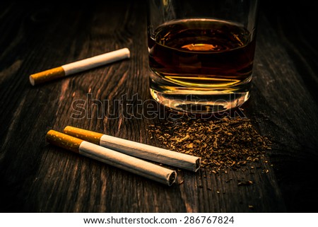 Glass of whiskey and three cigarettes with tobacco leaves on a wooden table. Focus on the tobacco, image vignetting and the orange-blue toning