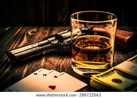 Glass of whiskey and playing cards with revolver and money on the wooden table. Angle view, image vignetting and the orange-blue toning, identification cards ace Russian letter